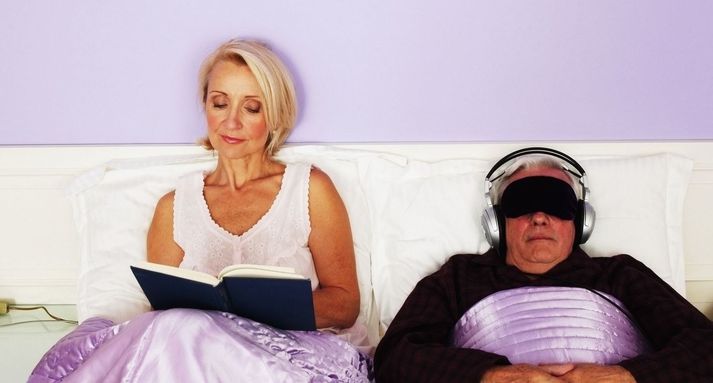 Couple in bed, man wearing eye mask and headphones, woman reading