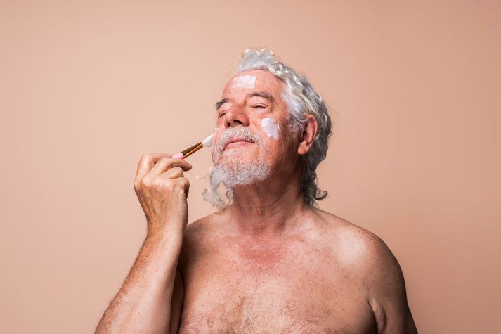 Senior man with eyes closed applying moisturizer using brush against beige background